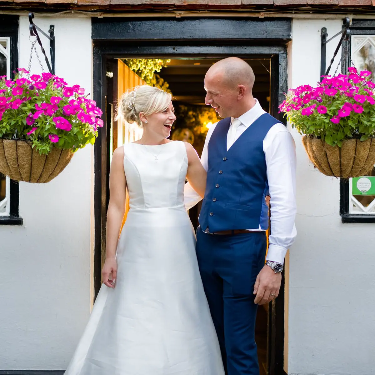 TJP_2018_09_08_00553_Creative_Wedding_Photographer A bride in a white gown and a groom in a blue vest and white shirt stand smiling at each other in a doorway, framed by hanging baskets of bright pink flowers—a perfect moment of creative wedding photography style.