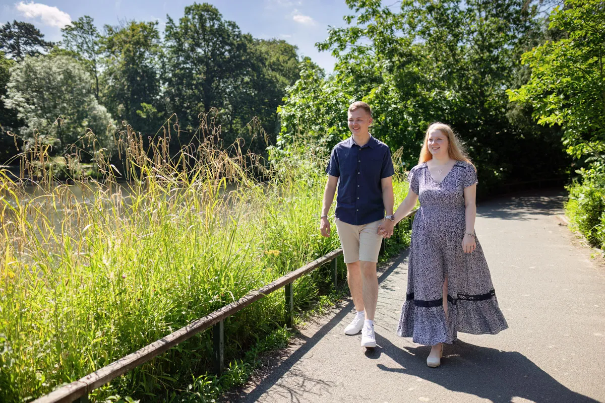 TJP_2024_06_27_0007 Strolling hand in hand through Kelsey Park's sunlit path, surrounded by greenery and tall grasses, the couple radiates happiness and relaxation. With a clear sky above, their engagement session captures her patterned dress and his blue shirt and shorts in perfect harmony.