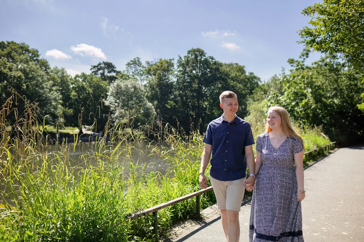 TJP_2024_06_27_0011 A couple enjoys an engagement session, holding hands as they walk along a sunlit path beside a lake in Kelsey Park. Surrounded by lush green foliage and trees under a clear blue sky, the woman dazzles in a patterned dress while the man is casually dressed in a blue shirt and shorts.