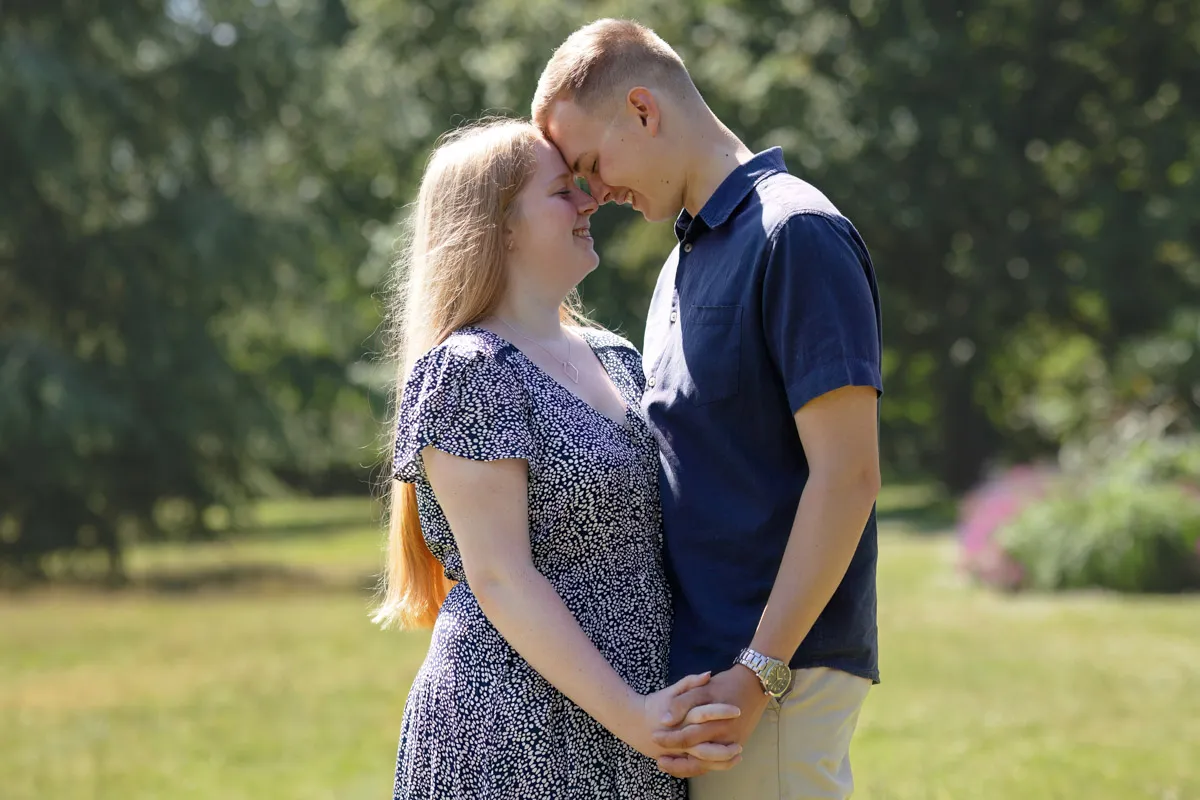 TJP_2024_06_27_0021 In the sunlit embrace of Kelsey Park, a couple stands closely, their foreheads touching and hands clasped—a tender moment captured during their engagement session. The woman dons a patterned dress, and the man sports a navy shirt with beige pants against the blurred green backdrop.