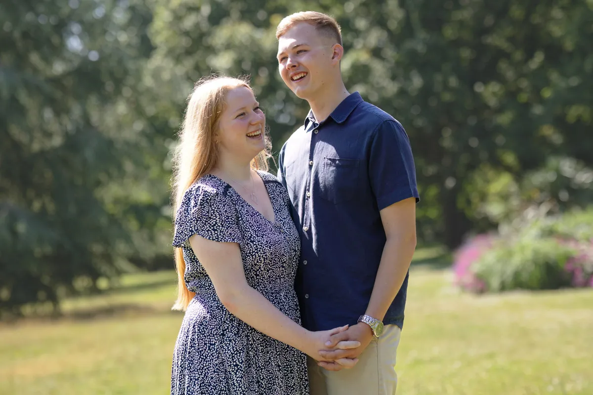 TJP_2024_06_27_0023 A couple stands outdoors in Kelsey Park during their engagement session, holding hands and smiling at each other. The woman is wearing a patterned dress, and the man is in a blue shirt and beige pants. They are standing on grass with trees and flowers blurred in the background.