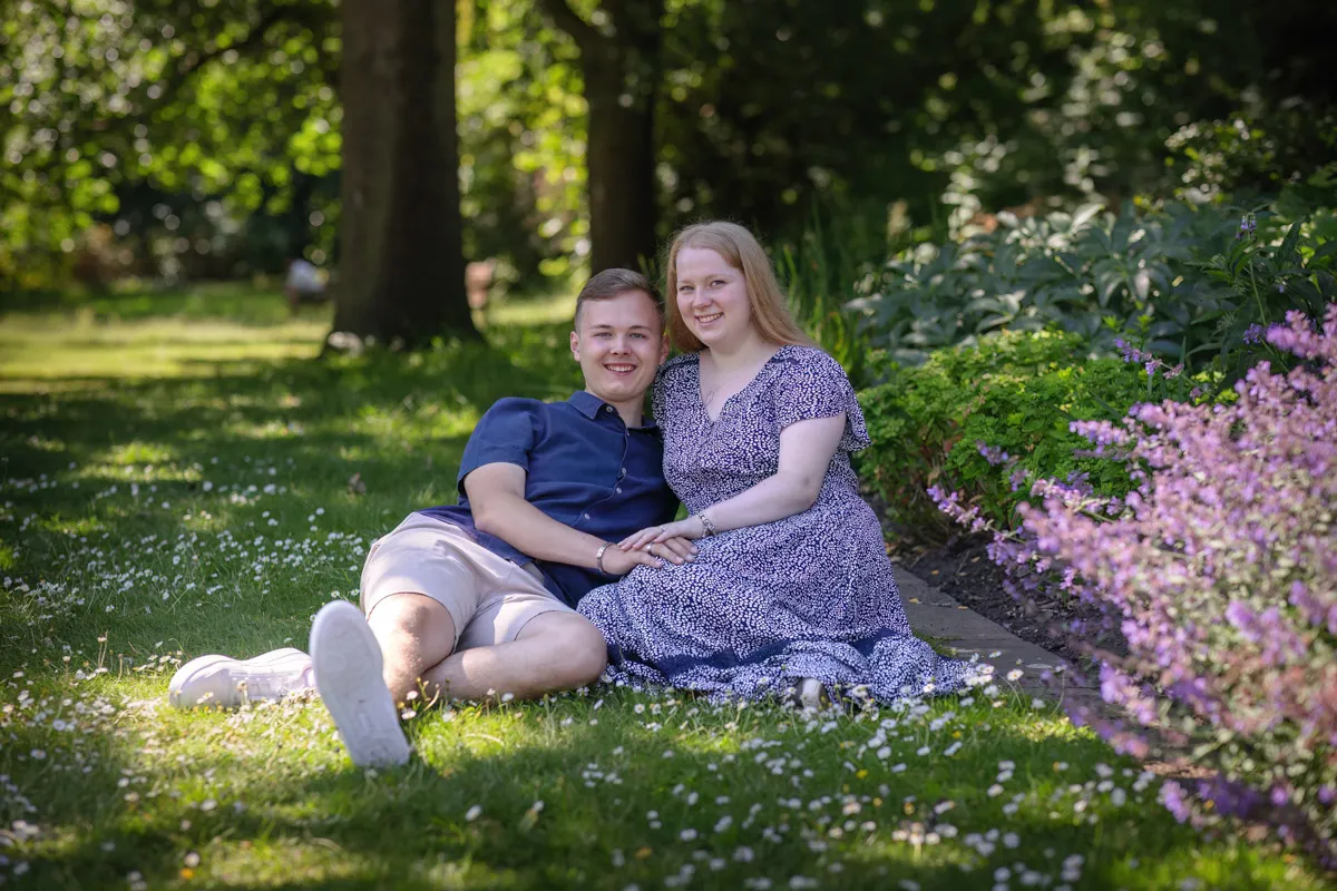 TJP_2024_06_27_0024 A couple is sitting on the grass in sunlit Kelsey Park, surrounded by lush greenery and purple flowers. They're leaning against each other, smiling, and holding hands during their intimate engagement session. Sunlight filters through the trees, creating a peaceful backdrop.