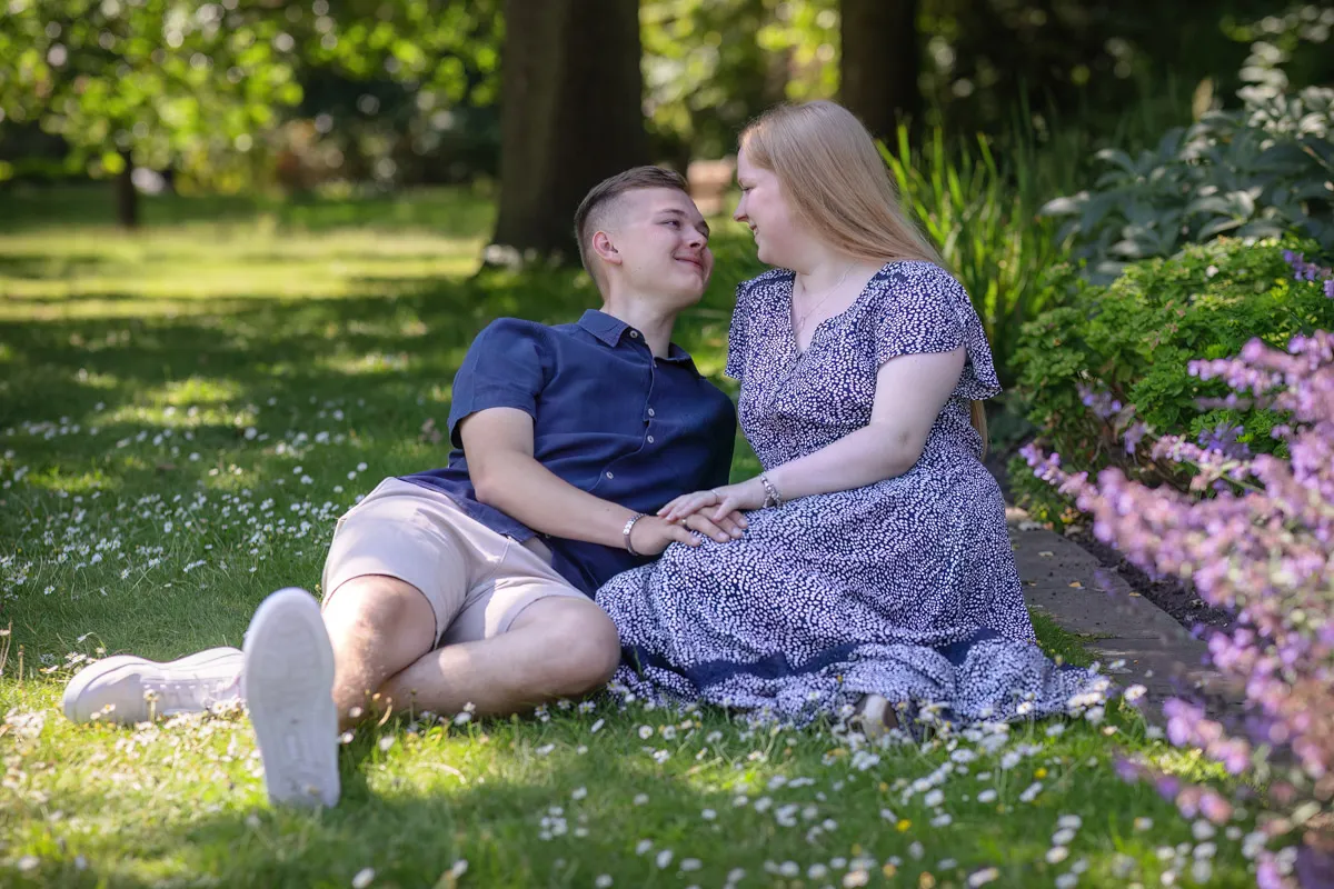 TJP_2024_06_27_0025 A couple sits on the grass in Kelsey Park, smiling at each other during their engagement session. The man wears a navy shirt and light shorts, while the woman is in a blue patterned dress. They hold hands amidst greenery and flowers, with sun filtering through the trees.