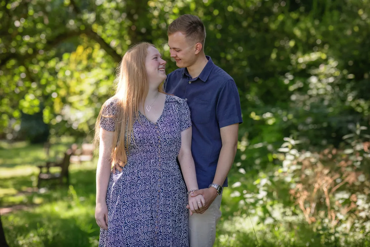 TJP_2024_06_27_0033 In a lush, green expanse of Kelsey Park, a couple stands hand in hand, gazing into each other's eyes. The woman wears a blue patterned dress, and the man is in a navy shirt and beige pants. Sunlight filters through the trees, casting a serene glow on their engagement session.