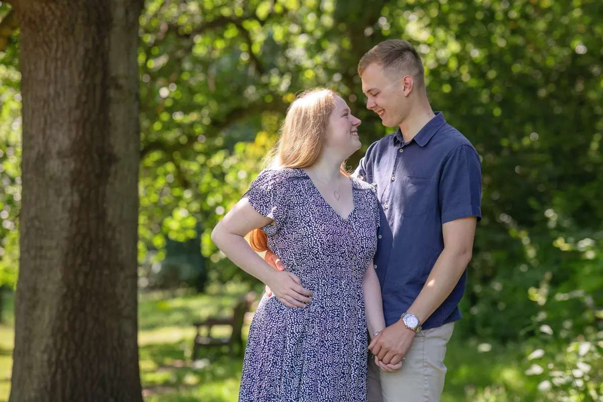 TJP_2024_06_27_0035 In a sunlit corner of Kelsey Park, a couple stands close under a tree, sharing smiles during their engagement session. The woman wears a patterned dress while the man sports a navy shirt and beige shorts. Bright green foliage and a bench grace the background.