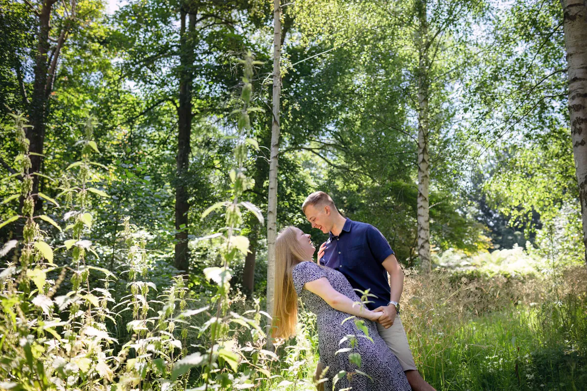 TJP_2024_06_27_0045 A couple enjoys a joyful moment during their engagement session in a sunlit, lush forest at Kelsey Park. The woman in a patterned dress leans back as the man in a blue shirt gently supports her, surrounded by tall grass and trees that create a serene atmosphere.