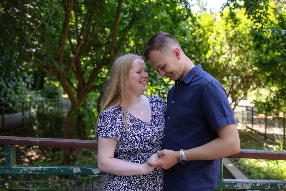 TJP_2024_06_27_0049 In the serene embrace of Kelsey Park, a couple stands closely together, smiling at each other. During their engagement session, they hold hands as sunlight dances through the trees; she in her patterned dress and he in his navy shirt.