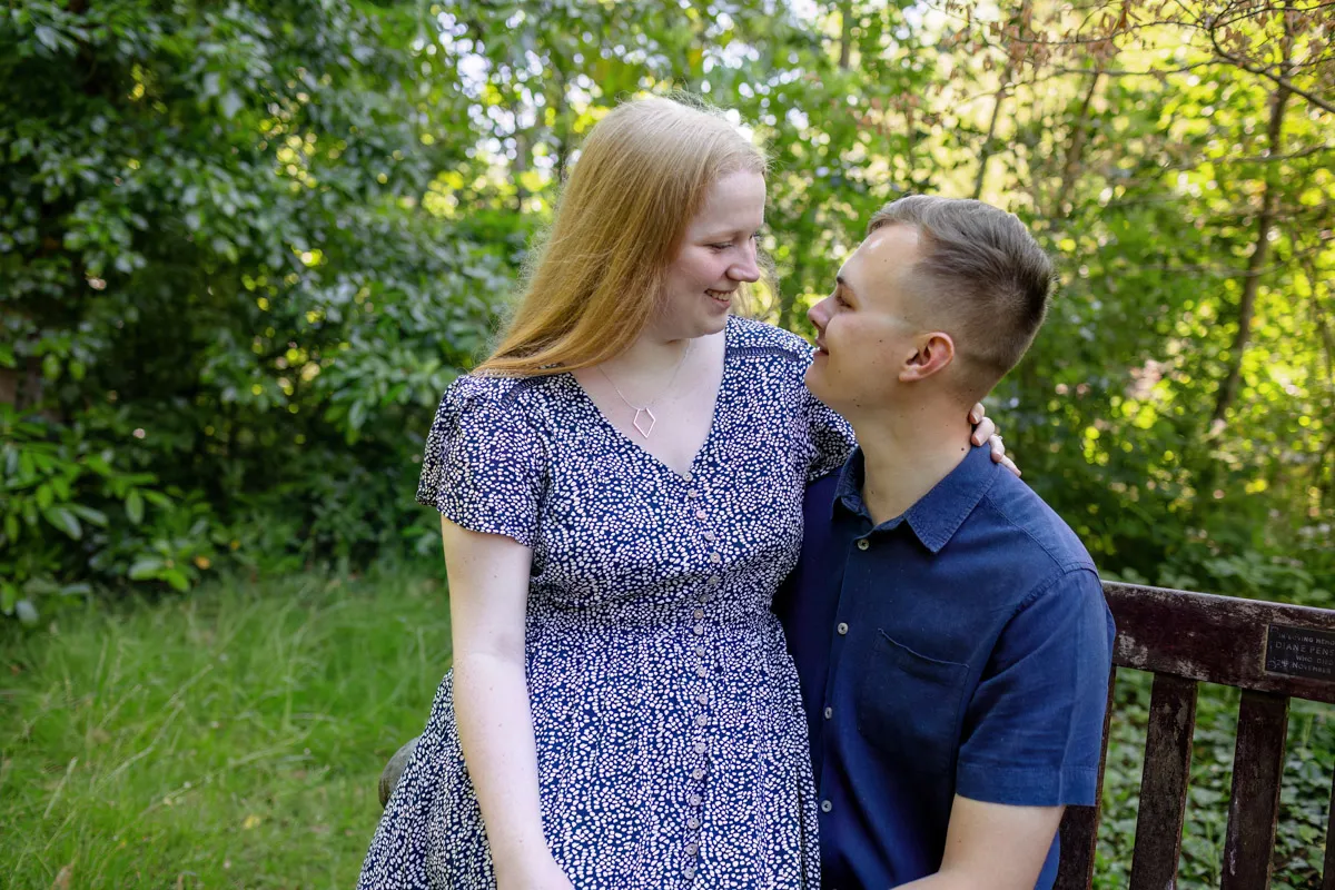 TJP_2024_06_27_0051 In Kelsey Park, an engagement session unfolds as a couple sits on a wooden bench. The woman, in a floral dress, smiles at the man in a navy shirt. They both appear content, surrounded by lush greenery and dappled sunlight.