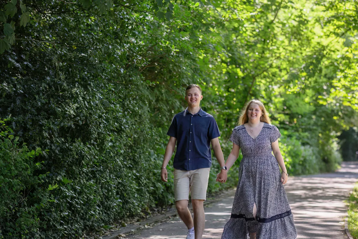 TJP_2024_06_27_0053 A young couple, possibly on their engagement session, holds hands as they stroll down a sunlit path in Kelsey Park. Surrounded by lush green trees and foliage, the man wears a dark shirt and shorts, while the woman is in a patterned dress. Both smile brightly and look ahead.