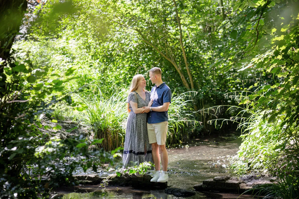 TJP_2024_06_27_0063 A couple stands together on stones in a creek at Kelsey Park, surrounded by lush greenery. During this enchanting engagement session, the woman wears a long dress and the man sports a blue shirt and shorts. Sunlight filters through the trees, creating a serene and romantic atmosphere.