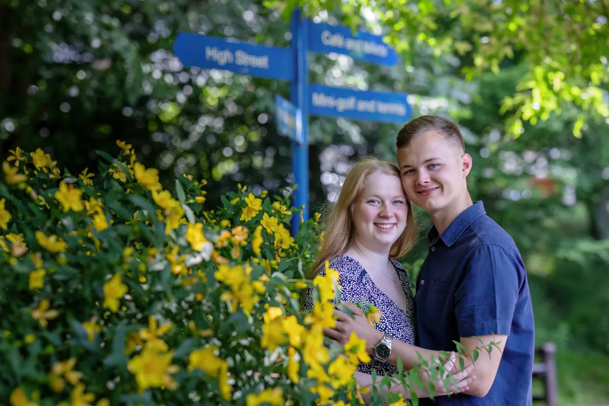 TJP_2024_06_27_0072 A couple stands closely, smiling amid lush greenery and vibrant yellow flowers at Kelsey Park. They are near a blue signpost with blurred text. Sunlight filters through the trees, adding a serene, romantic touch to this beautiful engagement session.