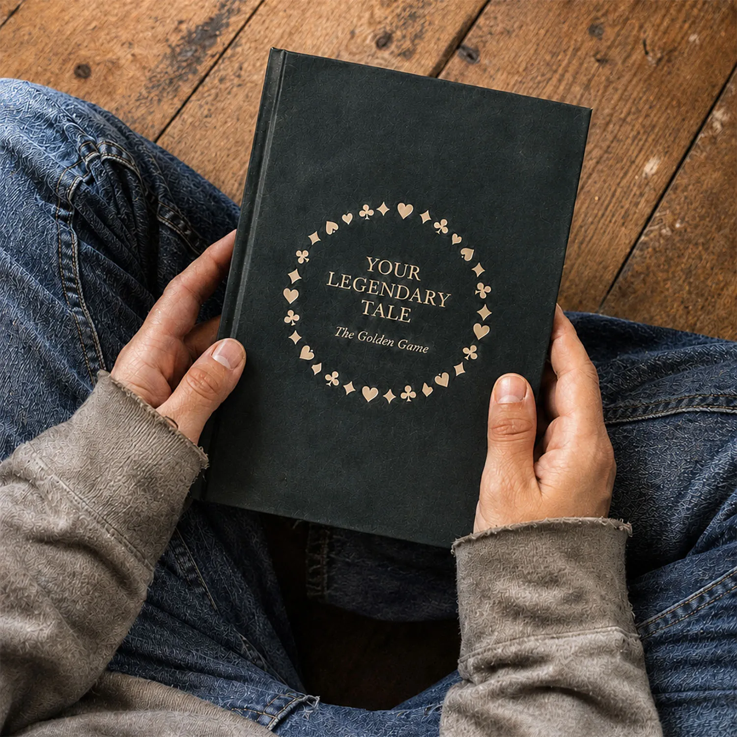 A person in jeans and a grey jumper sits on a wooden floor, holding "Your Legendary Tale: The Golden Game," a dark green book with gold card suits circling the title on the cover.