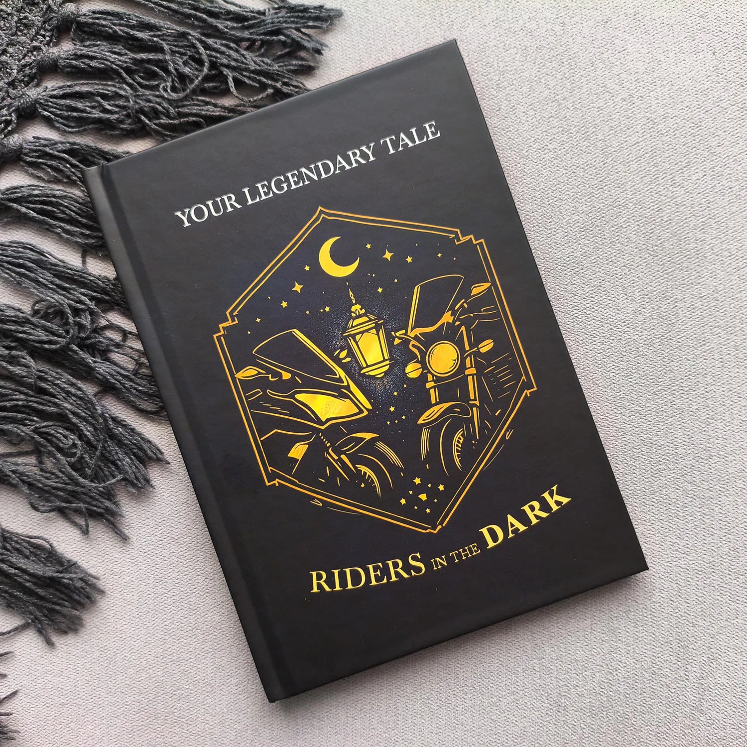 A black hardback book titled "Your Legendary Tale: Riders in the Dark" features a gold illustration of motorbikes riding under a crescent moon and stars, with a lantern and cityscape highlighting the adventure within Your Legendary Tale.