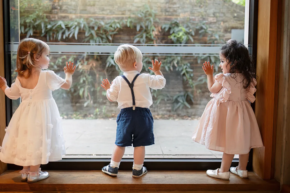 01_TJP_2025_08_16_0579 Three young children dressed in formal clothes stand on a windowsill, their hands pressed against the glass as they gaze outside—a charming moment captured during Pitzhanger Manor Wedding Photography.