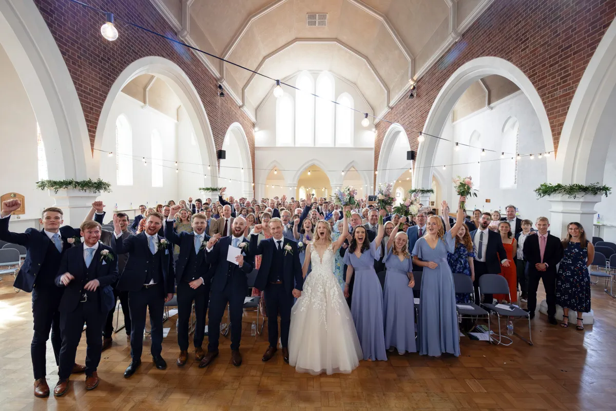 01_TJP_2025_08_30_0349 A large wedding party stands together in a bright, airy church hall. The Octagon Barn Norwich Wedding Photographer captures the bride and groom at the centre, surrounded by bridesmaids in blue dresses, groomsmen in suits, and many smiling guests.