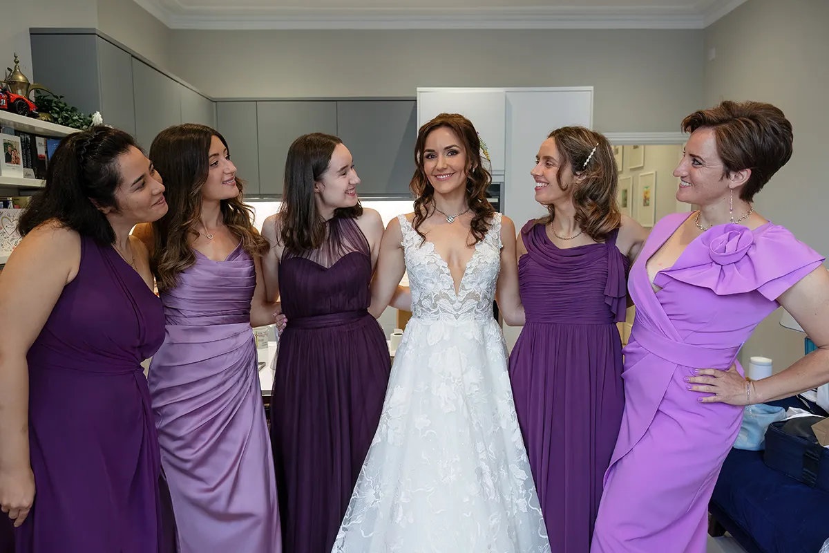02_TJP_2025_08_16_0015 A bride in a white dress stands smiling with five bridesmaids in matching shades of purple, all posing together happily in a bright room—capturing the joy of Pitzhanger Manor Wedding Photography.
