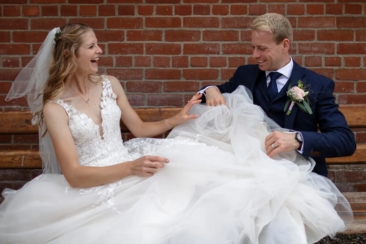 02_TJP_2025_08_30_0576 A bride in a white dress and veil laughs joyfully beside her smiling groom in a dark suit, both looking relaxed and happy against a red brick wall—captured by Octagon Barn Norwich Wedding Photographer.