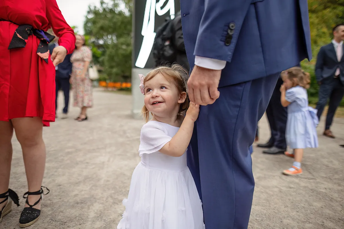 03_TJP_2025_08_16_0022 A young girl in a white dress smiles up at an adult in a blue suit, holding onto their leg. Other guests mingle in the background at an outdoor event, capturing the joy of Pitzhanger Manor Wedding Photography.