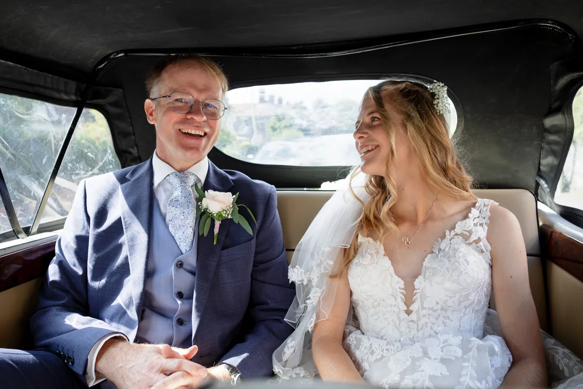 03_TJP_2025_08_30_0103 A bride in a white lace dress and veil sits beside a man in a blue suit with a buttonhole inside a car. They are smiling at each other, appearing happy and relaxed, as sunlight streams through the window—captured by Octagon Barn Norwich Wedding Photographer.