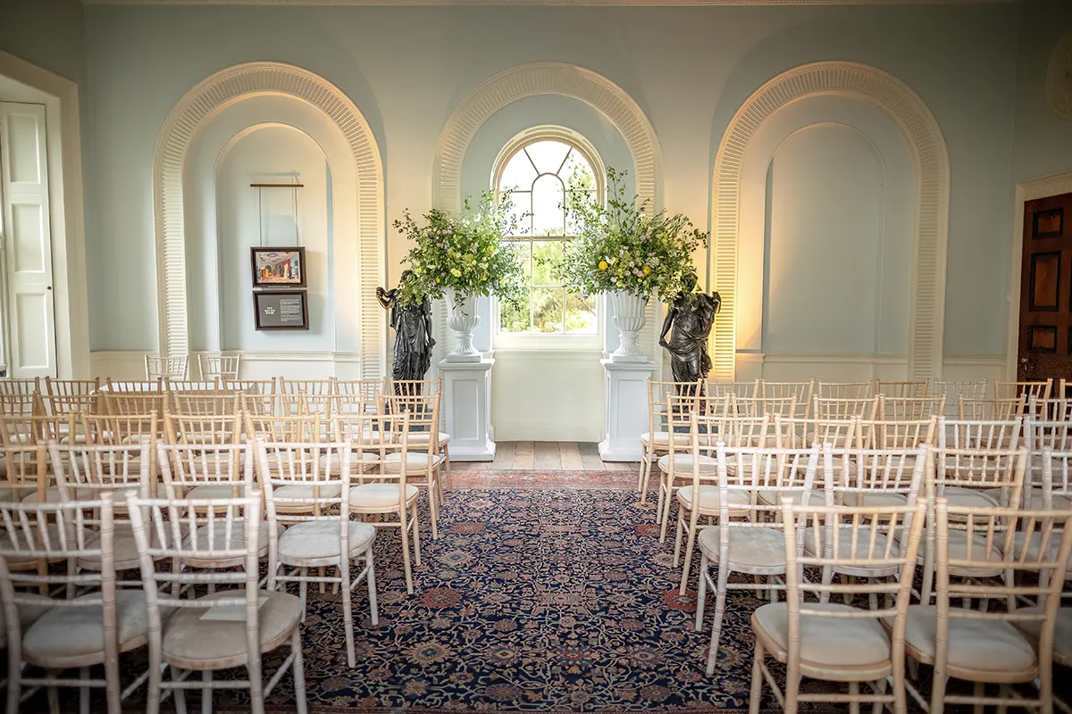 04_TJP_2025_08_16_0028 Rows of empty wooden chairs face a window framed by tall floral arrangements in a bright, elegant room with arched alcoves—a perfect Pitzhanger Manor Wedding Photography setting, with a patterned rug and soft lighting ready for the ceremony.