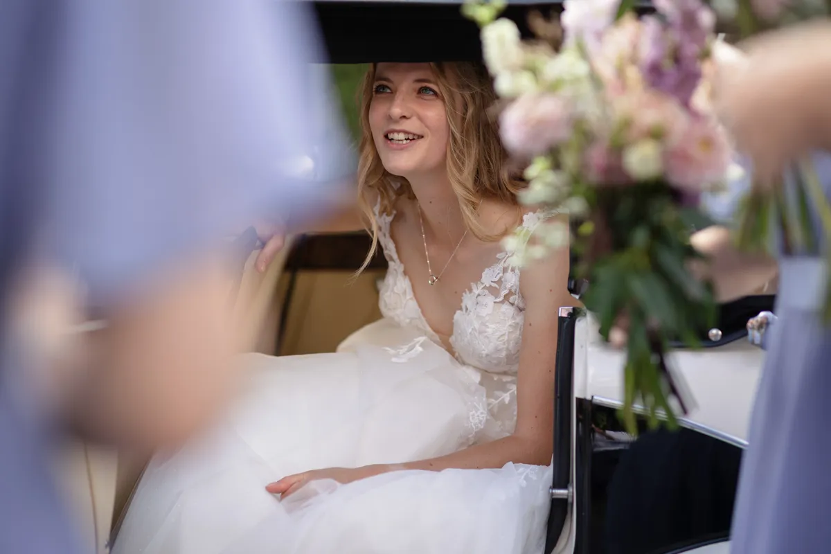 04_TJP_2025_08_30_0111 A smiling bride in a white lace wedding dress sits inside a car, looking out. Softly blurred flowers and people are visible in the foreground, beautifully captured by Octagon Barn Norwich Wedding Photographer.