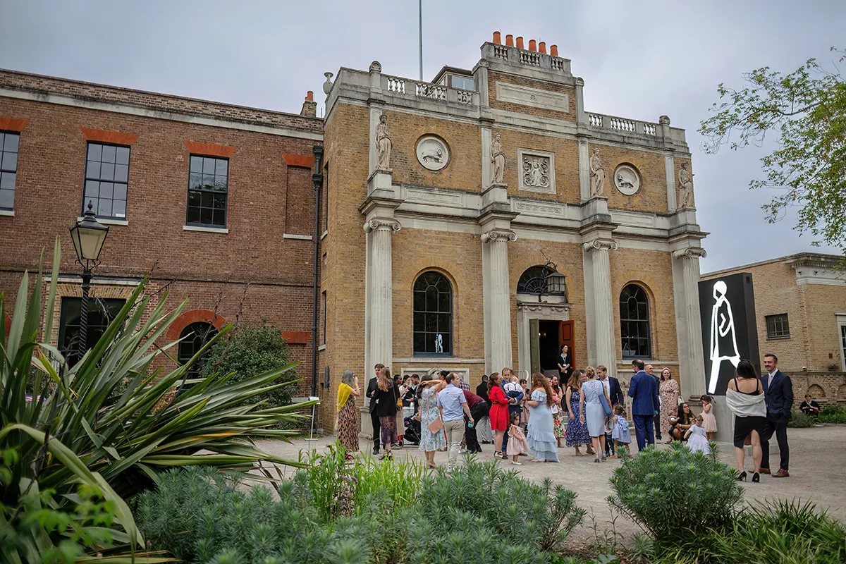 05_TJP_2025_08_16_0050 A group of people gather outside Pitzhanger Manor, its tall columns and classical statues providing a stunning backdrop for wedding photography. Lush greenery frames the historic scene beneath a cloudy sky.