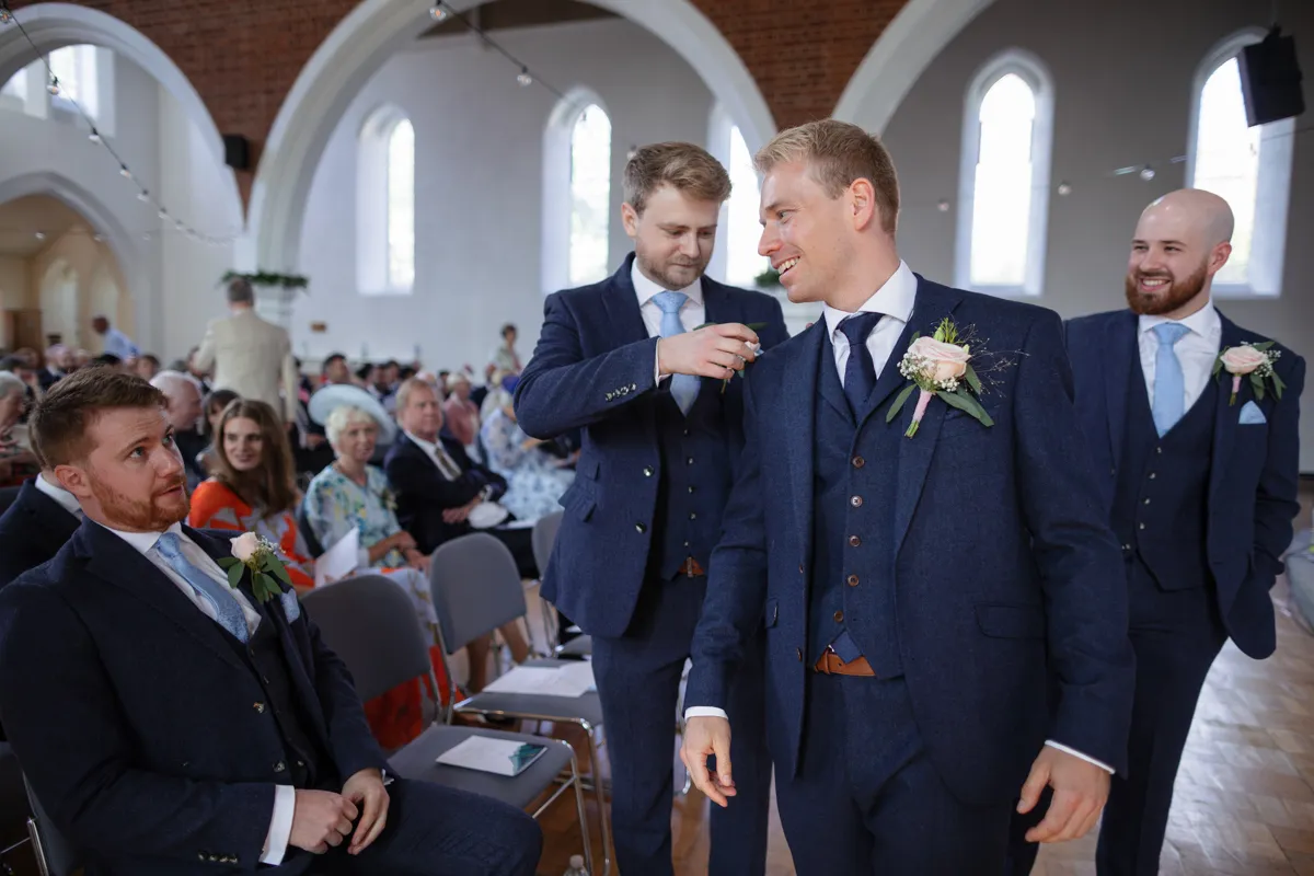 05_TJP_2025_08_30_0120 Four men in navy suits and light blue ties stand inside a bright church. One pins a buttonhole on another as guests watch. The cheerful yet formal atmosphere is perfect for an Octagon Barn Norwich Wedding Photographer to capture pre-ceremony moments.