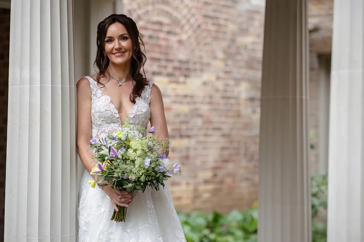 06_TJP_2025_08_16_0075 A smiling bride in a white lace dress holds a bouquet of flowers while standing between tall columns, with a brick wall and greenery in the background—capturing the elegance of Pitzhanger Manor Wedding Photography.