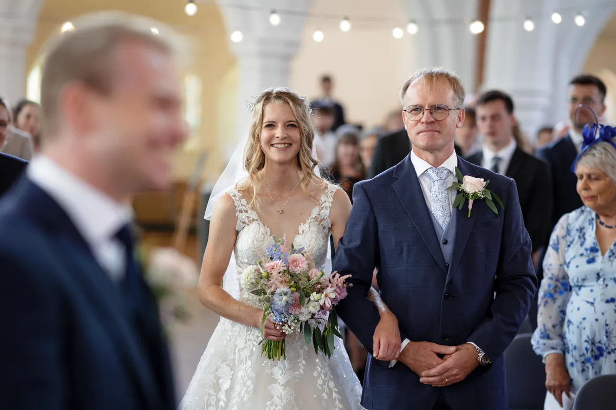 06_TJP_2025_08_30_0157 A bride in a white dress, holding a bouquet, walks down the aisle arm-in-arm with an older man in a suit. Captured by Octagon Barn Norwich Wedding Photographer, they smile as guests watch beneath blurred lights and arches.