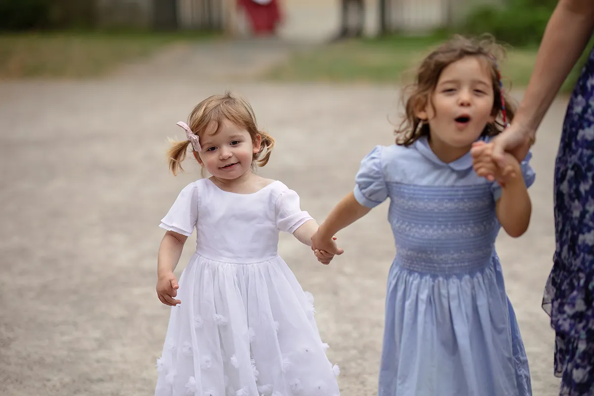 07_TJP_2025_08_16_0092 Two young girls in light dresses hold hands outdoors at a Pitzhanger Manor Wedding Photography session. The younger girl smiles softly, whilst the older girl appears to be singing or talking, an adult’s hand gently holding hers.