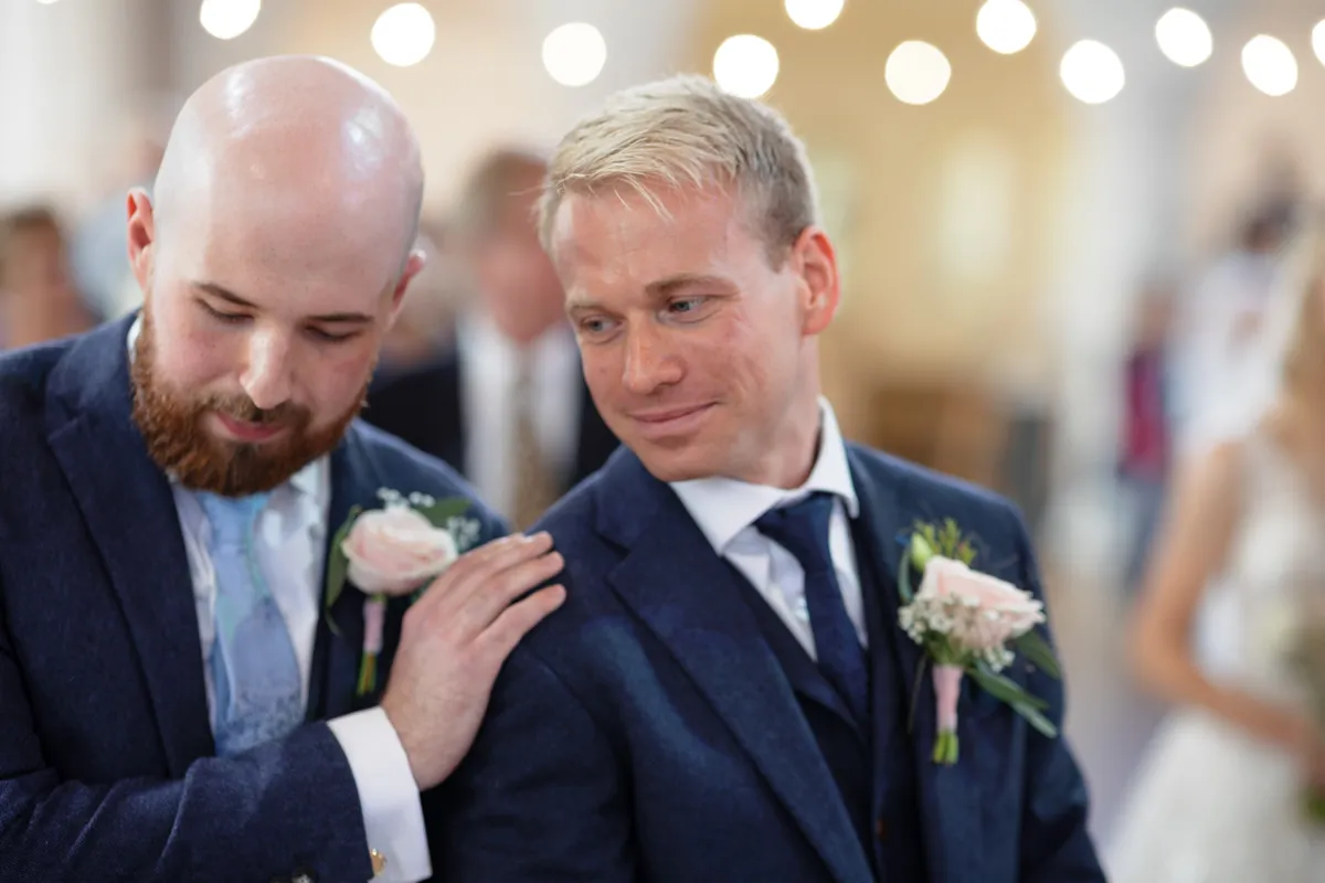 07_TJP_2025_08_30_0158 Two men in suits, both wearing buttonholes, stand close together at what appears to be a wedding. Captured by Octagon Barn Norwich Wedding Photographer, they share a warm moment under the soft glow of lights in the background.
