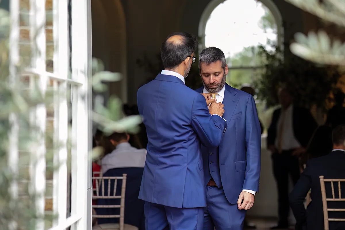 08_TJP_2025_08_16_0101 Two men in blue suits stand facing each other indoors; one adjusts the other’s buttonhole or tie. Captured in classic Pitzhanger Manor Wedding Photography style, people are seated behind them and greenery is visible through an open door.