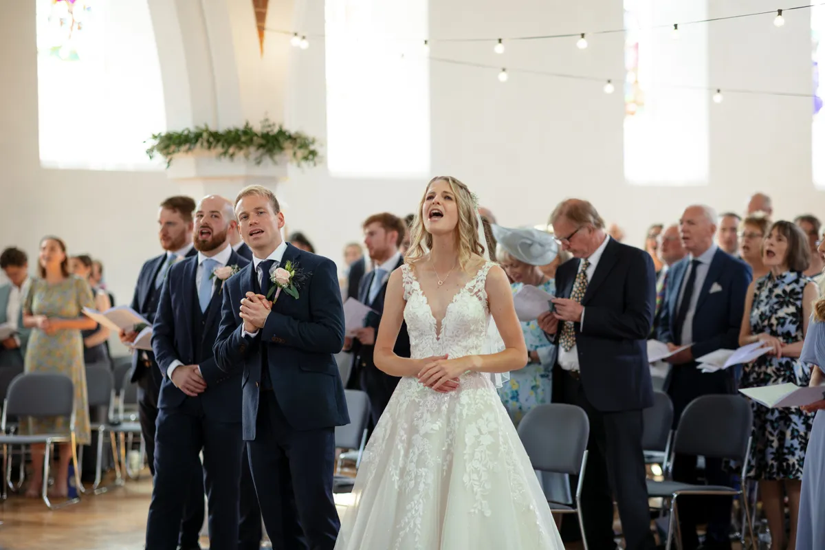 08_TJP_2025_08_30_0179 A bride in a white dress and guests, dressed formally, sing or speak during a wedding ceremony in the bright Octagon Barn Norwich venue, with fairy lights and large windows—perfect for any wedding photographer to capture.