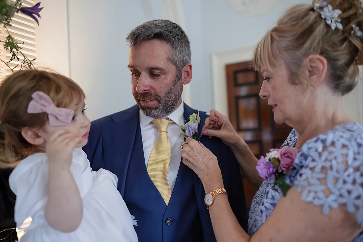 09_TJP_2025_08_16_0108 A woman adjusts a buttonhole on a man in a blue suit and yellow tie, while he holds a young girl in a white dress with a pink bow—capturing the candid moments of Pitzhanger Manor Wedding Photography as they prepare for a formal event indoors.