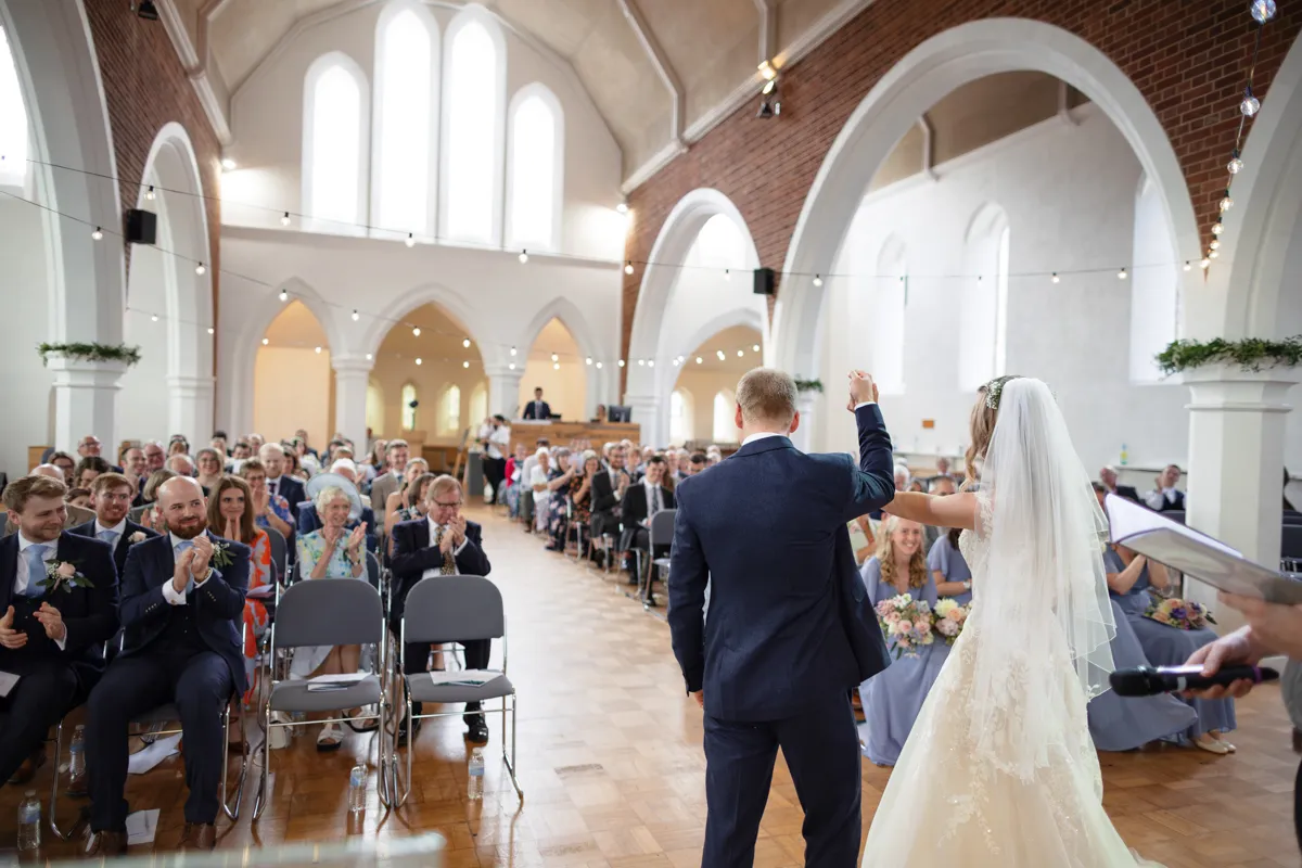 09_TJP_2025_08_30_0204 A bride and groom stand hand-in-hand at the front of a church filled with seated guests, celebrating their wedding ceremony as the audience applauds—captured beautifully by Octagon Barn Norwich Wedding Photographer.