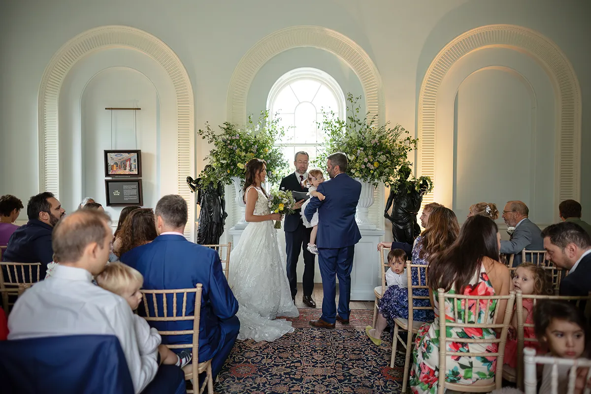 10_TJP_2025_08_16_0126 A bride and groom stand facing each other at their wedding ceremony in elegant Pitzhanger Manor, surrounded by guests. The groom holds a child, and floral arrangements with a large window complete this beautiful Pitzhanger Manor Wedding Photography moment.