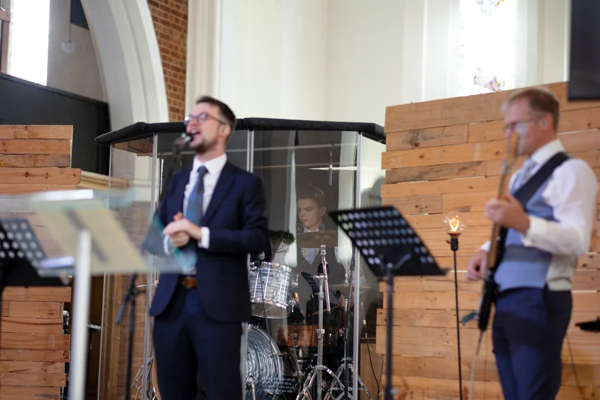 10_TJP_2025_08_30_0226 Three musicians perform indoors: a man sings into a microphone, another plays bass guitar, and a drummer is behind a drum kit inside a transparent enclosure. The wooden panels and tall windows evoke the charm of an Octagon Barn Norwich Wedding Photographer’s setting.