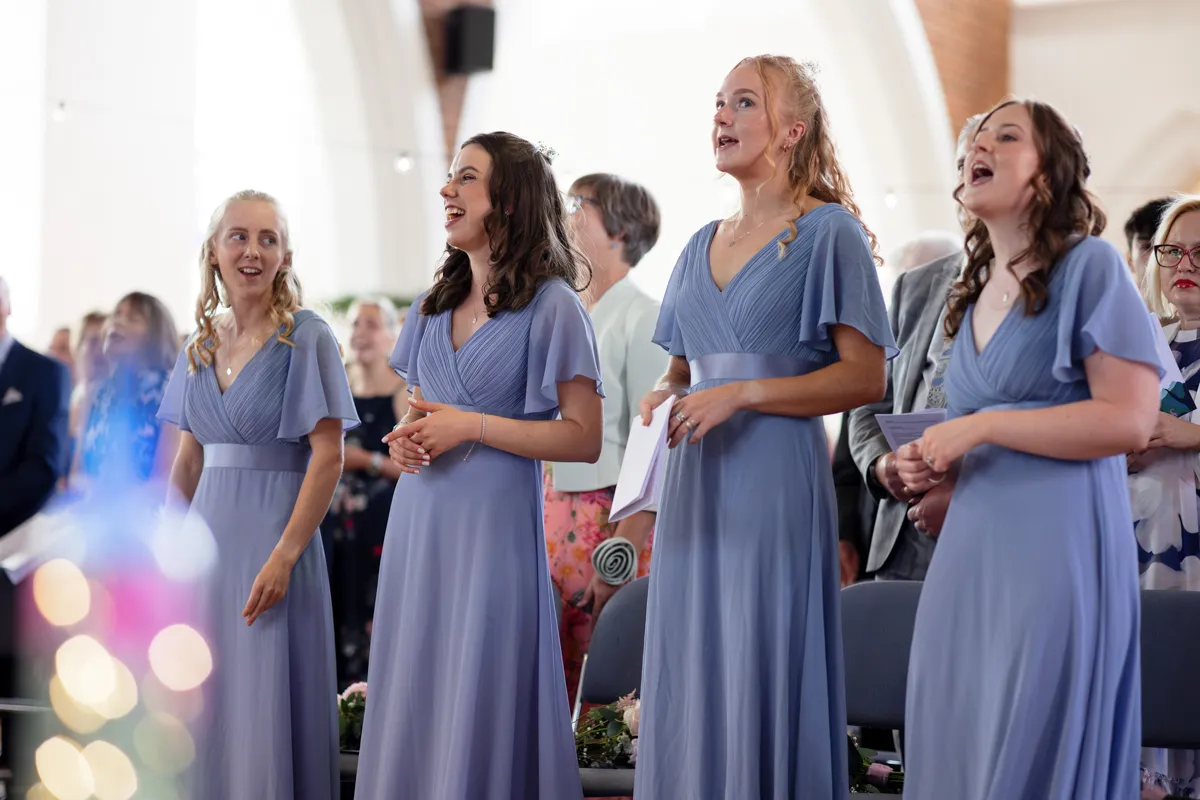 11_TJP_2025_08_30_0315 Four women in matching light blue dresses sing together in a bright indoor setting, possibly a church. Other guests look on joyfully, capturing a celebratory moment perfect for an Octagon Barn Norwich Wedding Photographer.