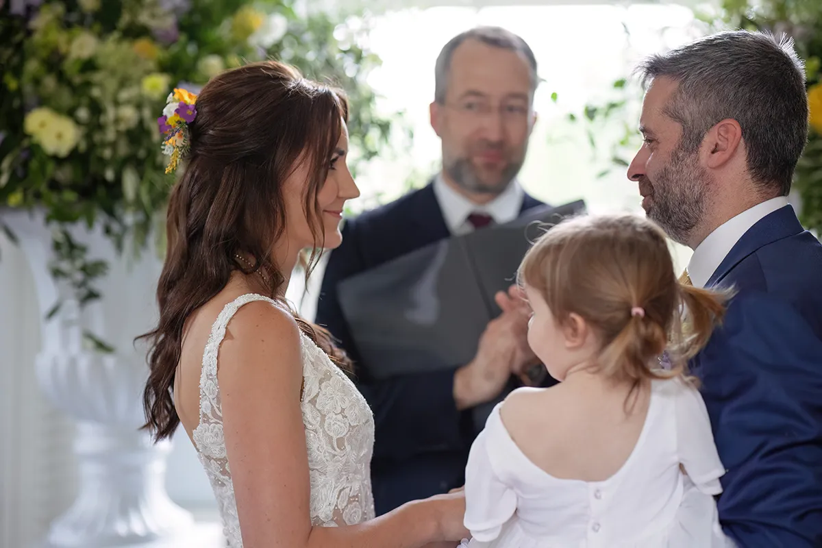 12_TJP_2025_08_16_0141 A bride and groom face each other, smiling, whilst holding a young girl during their Pitzhanger Manor Wedding Photography session. An officiant stands behind them, and flowers decorate the background.