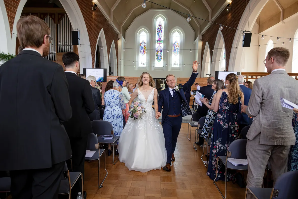 12_TJP_2025_08_30_0327 A bride and groom walk down the aisle, smiling and holding hands, as wedding guests stand and celebrate in a bright church with stained glass windows—captured beautifully by Octagon Barn Norwich Wedding Photographer.