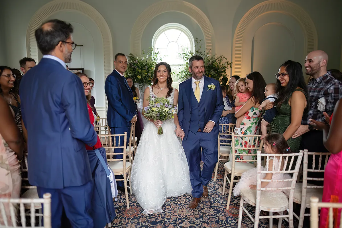13_TJP_2025_08_16_0178 A bride and groom smile as they walk down the aisle at their Pitzhanger Manor Wedding Photography session, surrounded by happy guests. The bride carries a bouquet in her white dress; the groom wears a blue suit with a yellow tie.