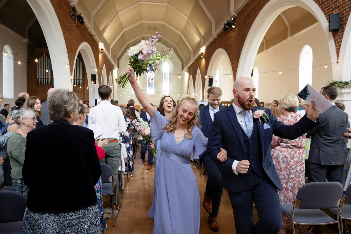 13_TJP_2025_08_30_0334 Two joyful wedding guests walk arm-in-arm down the aisle, beaming and celebrating. The woman in a lavender dress lifts her bouquet as the man in a suit holds papers. Captured by Octagon Barn Norwich Wedding Photographer, guests applaud along the aisle.