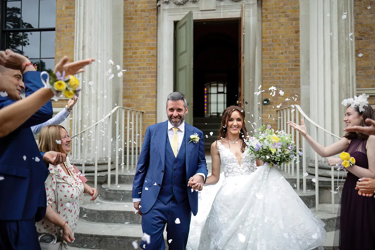 14_TJP_2025_08_16_0190 A bride and groom walk down steps outside a building as guests throw flower petals to celebrate. Captured in Pitzhanger Manor Wedding Photography style, the groom wears a blue suit whilst the bride holds her bouquet, both smiling happily.