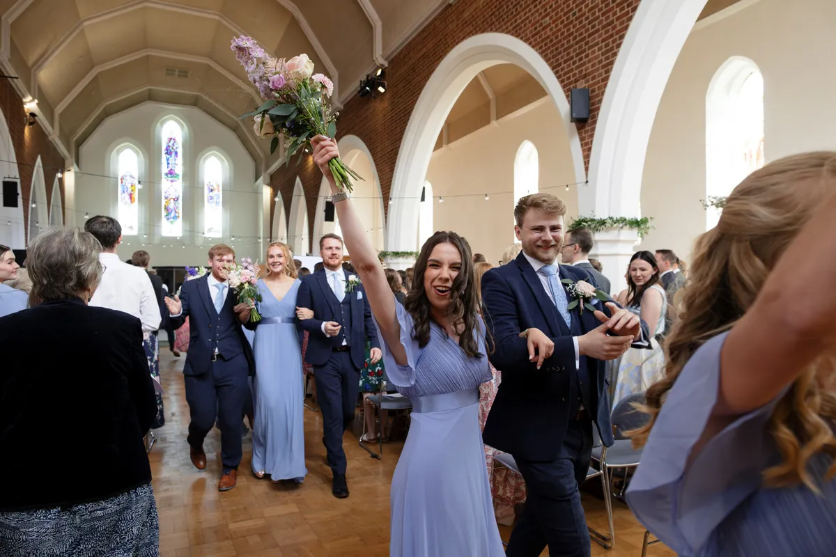 14_TJP_2025_08_30_0336 A joyful wedding party exits down the aisle of a bright church; bridesmaids in light blue dresses and groomsmen in suits smile, while one bridesmaid raises a bouquet—captured perfectly by Octagon Barn Norwich Wedding Photographer.