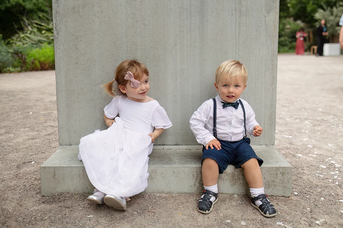 15_TJP_2025_08_16_0200 Two young children dressed in formal clothes sit on the ground against a concrete wall outdoors—a charming moment from a Pitzhanger Manor Wedding Photography session. The girl wears a white dress and bow; the boy sports braces, shorts, and a bow tie.