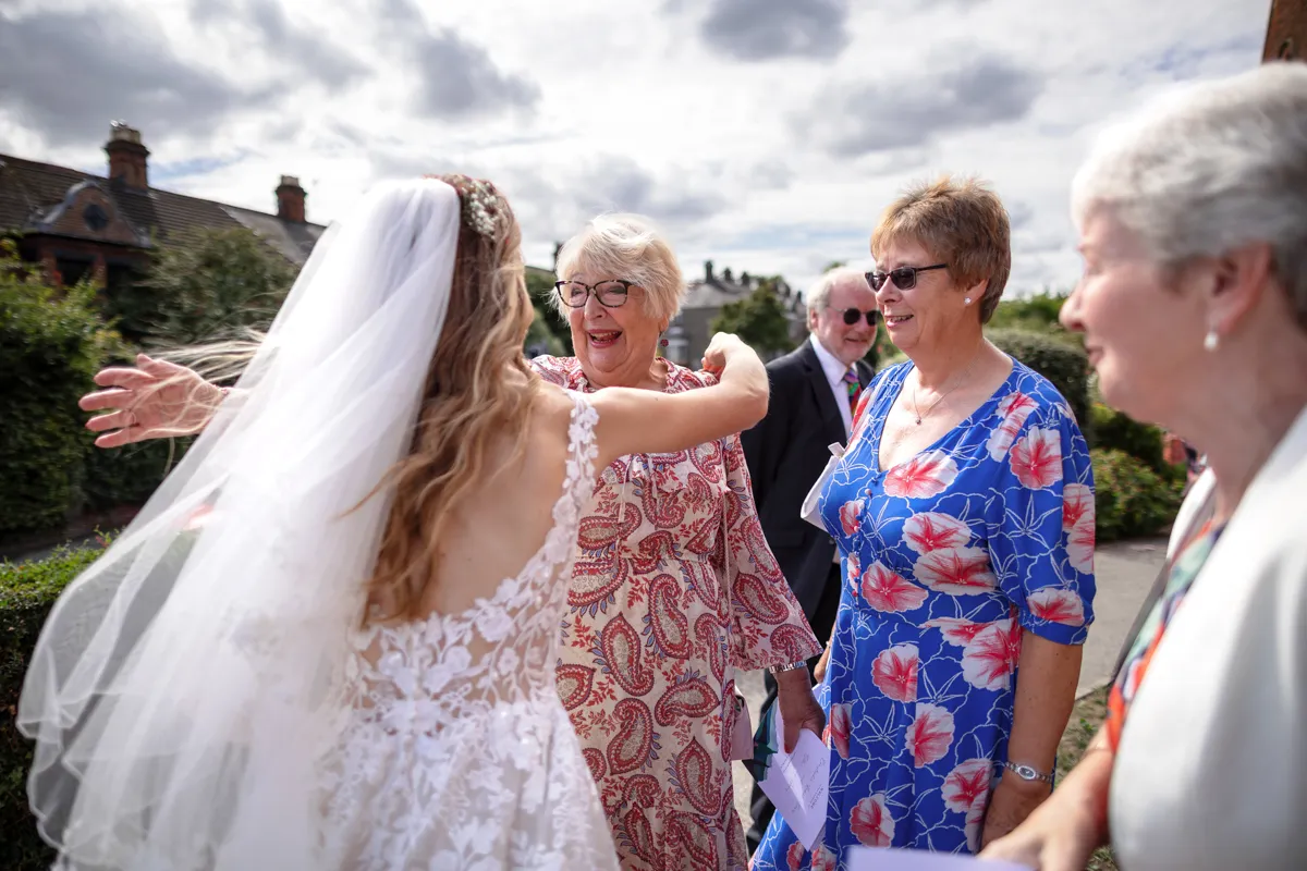 15_TJP_2025_08_30_0382 A bride in a white lace dress and veil joyfully embraces an older woman outside the Octagon Barn, while two other women and a man, all in colourful attire, watch and smile under a partly cloudy sky—captured by your Norwich Wedding Photographer.