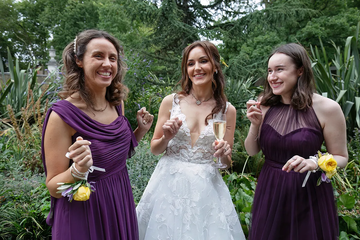 16_TJP_2025_08_16_0223 Three women stand outdoors, smiling and celebrating. The bride in a white wedding dress holds a champagne glass, flanked by bridesmaids in purple dresses with yellow flower corsages—capturing the joy of Pitzhanger Manor Wedding Photography. Green foliage fills the background.