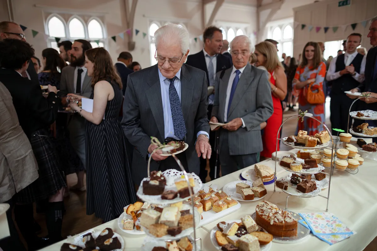 16_TJP_2025_08_30_0423 Elderly men in suits select cakes and pastries from a dessert table at a crowded indoor gathering, with people socialising in the background. The festive setting, captured by an Octagon Barn Norwich Wedding Photographer, features charming bunting decorations.