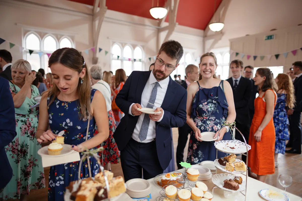 17_TJP_2025_08_30_0452 People in formal attire serve themselves cake and pastries from a dessert table at a social gathering in a bright, decorated hall with large windows and bunting—perfect for an Octagon Barn Norwich Wedding Photographer to capture.