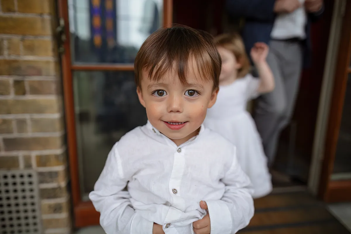 18_TJP_2025_08_16_0273 A young boy in a white shirt stands indoors, looking at the camera with a slight smile. Behind him, a girl in a white dress and an adult are partially visible in the softly lit Pitzhanger Manor Wedding Photography setting.